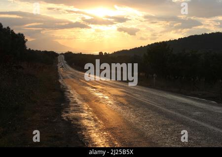 Strada asfaltata vuota con corsie bianche senza automobili o traffico. Cieli nuvolosi, montagne, alberi e l'alba all'orizzonte mentre il sole scende. Foto Stock