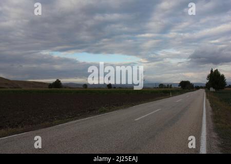 Strada asfaltata vuota con corsie bianche senza automobili o traffico. Cieli nuvolosi, montagne, alberi e l'alba all'orizzonte mentre il sole scende. Foto Stock