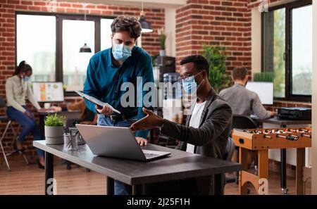 Diversi uomini d'affari che lavorano al progetto di marketing che analizza il grafico aziendale discutere idee di business in mattone muro startup ufficio. Squadra multietnica che indossa maschera medica contro covid19 Foto Stock