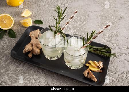 due bicchieri di limonata di zenzero fatta in casa su un vassoio scuro in ceramica con rosmarino, zenzero. fondo in cemento grigio Foto Stock