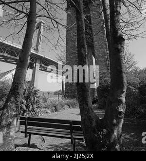 Vista in bianco e nero di una panca vuota in un parco appartato sotto il Royal Albert Bridge a Saltash, offre ai visitatori la vista del fiume Tamar e Th Foto Stock