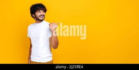 Foto panoramica di un ragazzo indiano o arabo dai capelli ricci positivo, guardando e puntando il dito di lato a uno spazio vuoto mock-up, mentre in piedi su sfondo arancione isolato, sorridente Foto Stock