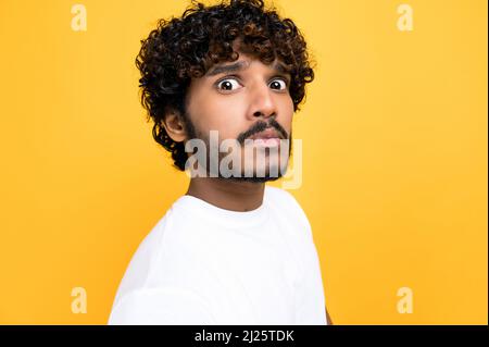 Primo piano di un ragazzo confuso indiano o arabo temuto con capelli ricci, guardando la fotocamera in incredulità, in piedi su un isolato sfondo arancione indossando una t-shirt bianca Foto Stock