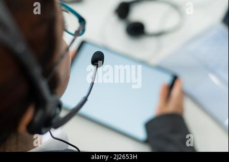 Vista posteriore di una donna in un auricolare che utilizza un tablet digitale mentre si siede alla scrivania. Operatore di servizio di supporto femminile al lavoro. Foto Stock