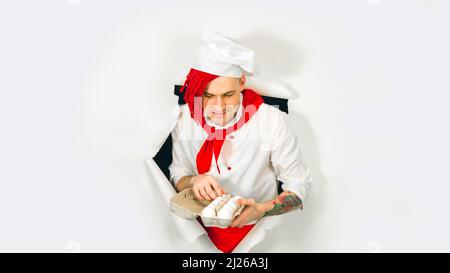 Un bell'uomo con lucchetti rossi lo tiene nelle sue uova di pollo in mano. Giovane cuoco sicuro in camicia bianca e cravatta rossa che tiene le uova di pollo, peeking th Foto Stock