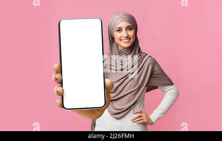 Felice giovane donna musulmana in hijab mostrando cellulare con schermo vuoto, dimostrando dispositivo con mockup Foto Stock