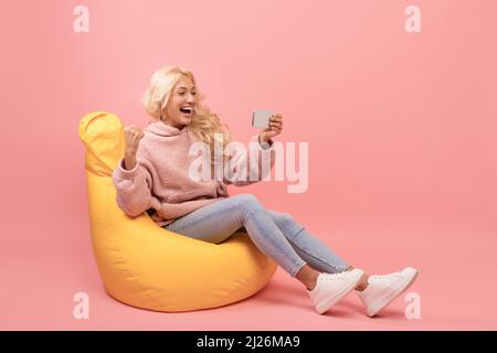 Felice signora tenere smartphone e gesturing sì nella gioia, leggere buone notizie, seduta in poltrona beanbag, sfondo rosa Foto Stock