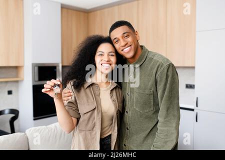 Coppia eccitata multirazziale in amore si muovono in un nuovo appartamento. Donna allegra nell'abbraccio del suo ragazzo che tiene le chiavi. Newlywed guardando la macchina fotografica e sorridere Foto Stock