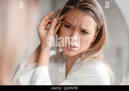 Rimozione dei capelli in eccesso dal viso. Giovane donna in sleepwear cazzo sopracciglia in bagno interno, closeup Foto Stock