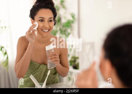 Bella giovane donna bruna che indossa un asciugamano dopo il bagno, rinfrescando la sua pelle con lozione o tonico vicino allo specchio a casa Foto Stock