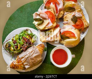 Selezione di sandwich, croissant e pane affettato con avocado, prosciutto, formaggio e verdure sul piatto, tazza di tè sul tavolo, primo piano. Foto Stock