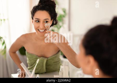 Bella donna millennial sorridente in tovagliolo utilizzando ciglia arricciatore vicino specchio a casa, copia spazio Foto Stock