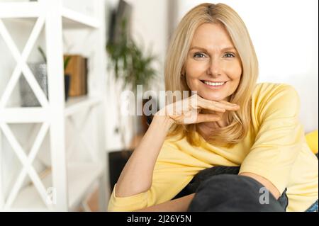 Ritratto di un affascinante uomo d'affari maturo di mezza età con le mani su un mento sorridente alla macchina fotografica, seduto nella casa moderna, indossando maglione giallo Foto Stock