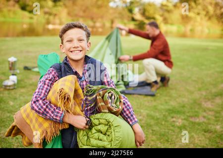 Questo viaggio in campeggio sarà impressionante. Ritratto di un ragazzo che tiene l'attrezzatura da campeggio con il padre sullo sfondo. Foto Stock