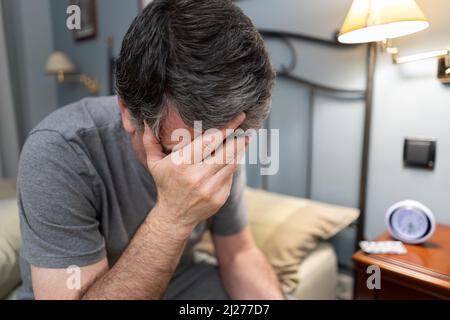 Uomo con insonnia seduto sul bordo del letto e con le mani sul viso. Foto Stock