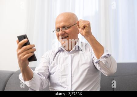 Eccitato anziano anziano pensionato uomo che guarda lo schermo del telefono, che celebra la vittoria in linea della lotteria o che ottiene il messaggio con le buone notizie. Emotivo maturo Foto Stock