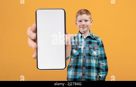 Ragazzo rosso sorridente che mostra un grande smartphone con schermo bianco vuoto alla fotocamera Foto Stock