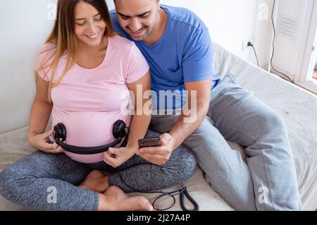 felice caucasica giovane donna incinta con cuffie sul pancino e marito che suona la musica con il telefono per il bambino, coppia incinta eterosessuale coccolare ba Foto Stock