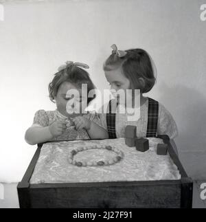 1961, storica, due giovani ragazze, sorelle, in piedi uno sgabello salotto, giocando con mattoni giocattolo o blocchi di legno e una collana di legno, Stockport, Manchester, Inghilterra, Regno Unito. Foto Stock