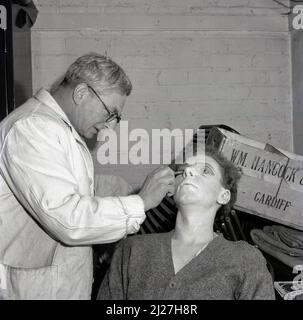 1950s, storico, un performer maschile che ha il make-up, occhio ombra applicato prima di una performance ai dipendenti della Steel Company of Wales, Abbey Works, Port Talbot, Wales, UK. Una cassa di legno di birrifici William Hancock & Co di Cardiff può essere visto riposare su alcune sedie dietro di loro. Foto Stock