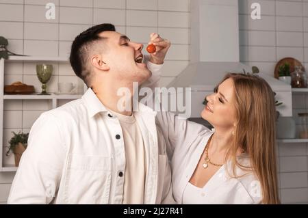 Donna che tratta, alimenta l'uomo. Moglie che mette il pomodoro ciliegia in bocca dell'uomo. Coppia di amore divertirsi in cucina a casa. Foto Stock