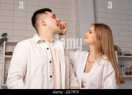 Donna che tratta, alimenta l'uomo. Moglie che mette il pomodoro ciliegia in bocca dell'uomo. Coppia di amore divertirsi in cucina a casa. Foto Stock