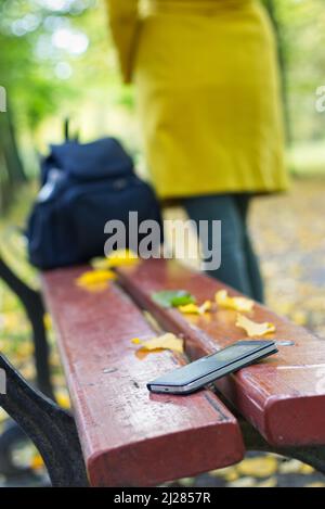 Smartphone dimenticato su una panca da parco. La donna sta lasciando da una panca dove ha perso il suo telefono cellulare. Foto Stock
