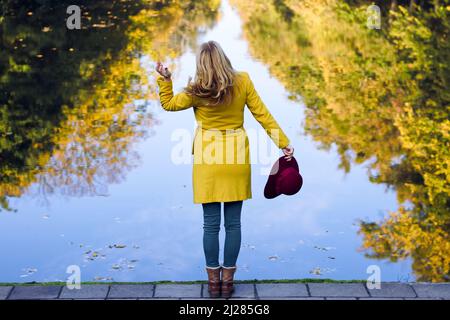 Donna capelli biondi con cappotto giallo e cappello rosso. Bella donna in piedi sul lago in autunno parco Foto Stock