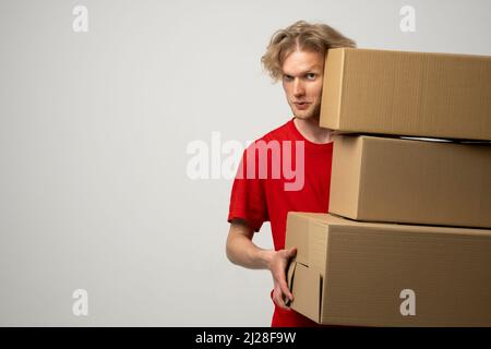 Giovane uomo di consegna in una uniforme rossa che tiene una pila di scatole di cartone. Corriere che consegna pacchi postali, pacchi su sfondo bianco studio. Foto Stock