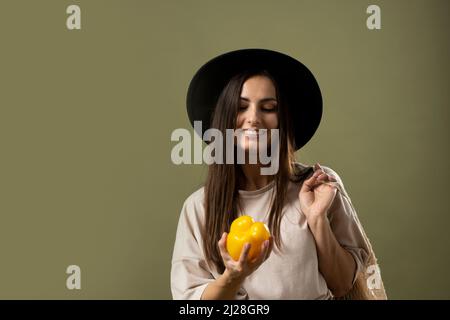 Donna eco-compatibile sorridente in t-shirt beige e cappello nero con sacchi eco di cotone riutilizzabili pieni di generi alimentari su una spalla e pepe giallo. Foto Stock