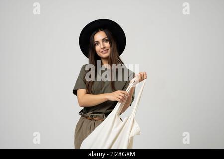 Felice donna bruna sorridente in cappello nero con borsa ecologica in cotone bianco sulla spalla. Ragazza che tiene in mano la borsa di prodotti tessili con verdure. Zero sprechi Foto Stock