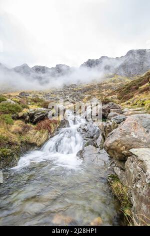 Bassa pov di un ruscello di babbling (che genera una piccola cascata) mentre l'acqua sta scorrendo giù una montagna nebbiosa, Snowdonia National Park, Galles del Nord, Regno Unito. Foto Stock