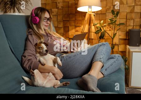 Ragazza attraente ascolta la musica e guarda i video clip su un tablet come un cane in un ambiente domestico al chiuso Foto Stock