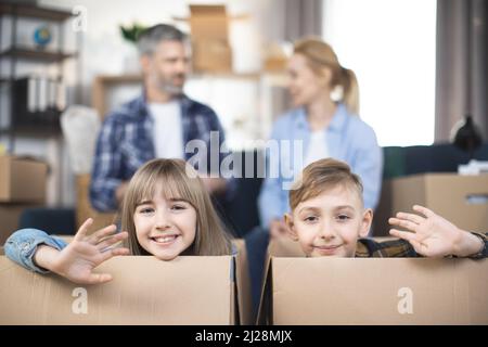 Due divertenti bambini sorridenti, ragazzo e ragazza, che guardano e sventolano la macchina fotografica dalla scatola di cartone mentre i loro genitori si siedono sul divano dietro. Felice famiglia godendo trasferimento in nuovo appartamento. Foto Stock