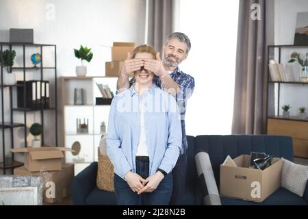 Felice bel bearded uomo che copre gli occhi della sua moglie affascinante eccitata mentre in piedi in nuovo appartamento alla moda dopo il trasferimento. Coppia allegra che si sposta a casa nuova Foto Stock