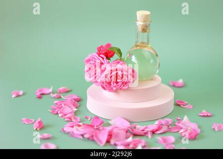 Olio essenziale di rosa in bottiglia di vetro in fiori di rosa in petali di rosa su un podio rosa su sfondo verde. Aromaterapia e cosmetici. Rosa naturale Foto Stock