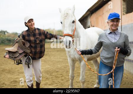 Donna asiatica che tiene la sella mentre donna più anziana rancher che conduce il cavallo all'aperto Foto Stock