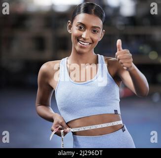Fai del tuo meglio e i risultati saranno mostrati. Ritratto di una giovane donna sportiva che mostra i pollici in su mentre misura la sua vita in una palestra. Foto Stock