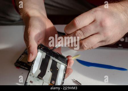 Repair of smartphones concept. A man unscrews the screws fixing the display from the board. Part of a series. Inside the room. Selective focus. Foto Stock