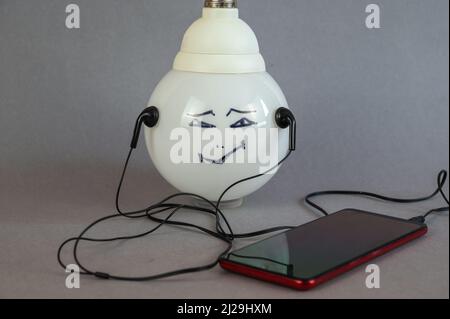 Una grande lampada elettrica bianca con il volto di un uomo dipinto su di essa. Uno smartphone e cuffie cablate. Composizione su sfondo grigio. Foto Stock