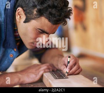 La prima volta. Scatto di un handyman focalizzato che misura un pezzo di legno mentre lavora nella sua officina. Foto Stock