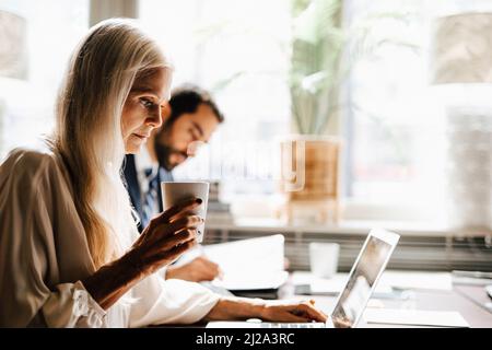 Donna d'affari matura che tiene la tazza di caffè usando il laptop da uomo d'affari alla scrivania in ufficio Foto Stock