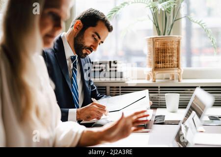 Uomo d'affari che analizza il documento da parte di donna d'affari che usa il laptop alla scrivania in ufficio Foto Stock