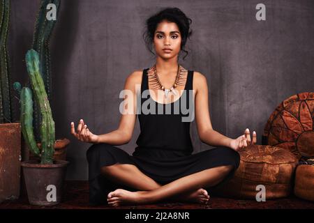 Meditando la magia. Studio ritratto di una bella giovane donna seduta in posizione loto accanto a un cactus e cuscini. Foto Stock