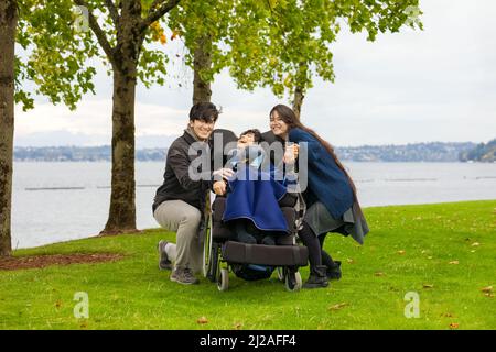 Giovane coppia biraciale che si piega accanto al ragazzo disabili in sedia a rotelle all'aperto al parco vicino al lago in giornata colma Foto Stock