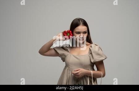 Ritratto di donna bruna affascinante in un abito beige con sacco eco in maglia di cotone e un peperone rosso tiene vicino ad un viso. Concetto di shopping senza rifiuti. Foto Stock