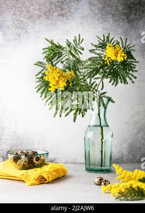 Bel bouquet di fiori d'argento o mimosa gialli in vaso di vetro turchese e piatto con uova di quaglia. Ancora vita. Foto Stock