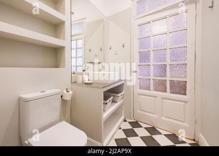 WC con lavabo incorporato con lavandino in porcellana bianca, porta in legno con vetri opachi e pavimenti piastrellati a scacchi Foto Stock