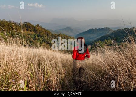 Una donna in manica lunga rossa con un cappello bianco cammina sulle praterie secche dorate di Hadubi, Chiang mai, Thailandia. Foto Stock