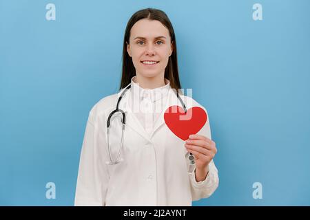 Ritratto di amichevole sorridente giovane medico in cappotto medico bianco professionale con stetoscopio che tiene rosso il cuore, guardando la macchina fotografica, posando l'isola Foto Stock